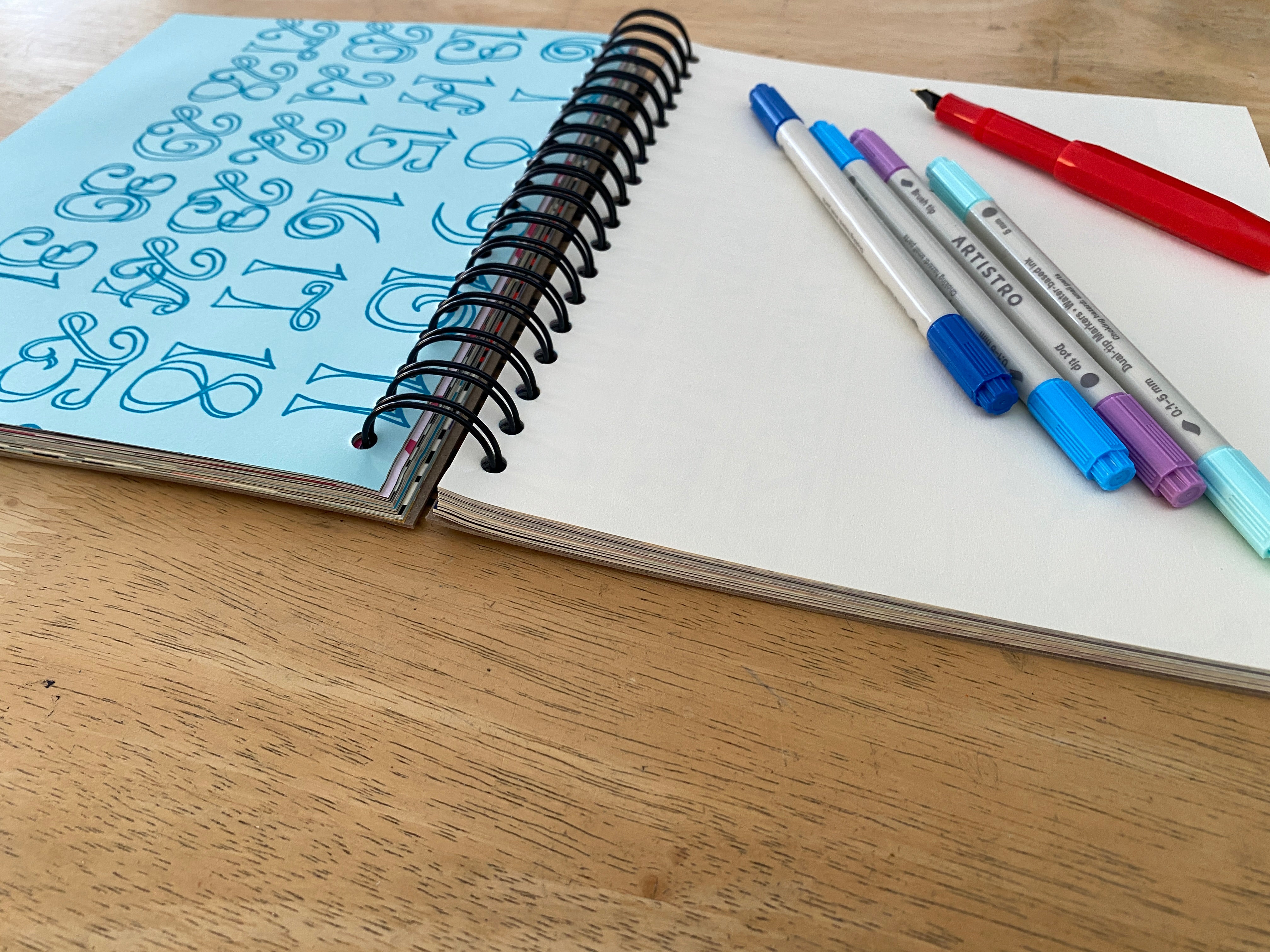 Open notebook with blue cover and black spiral binding, accompanied by colorful markers on a wooden surface.