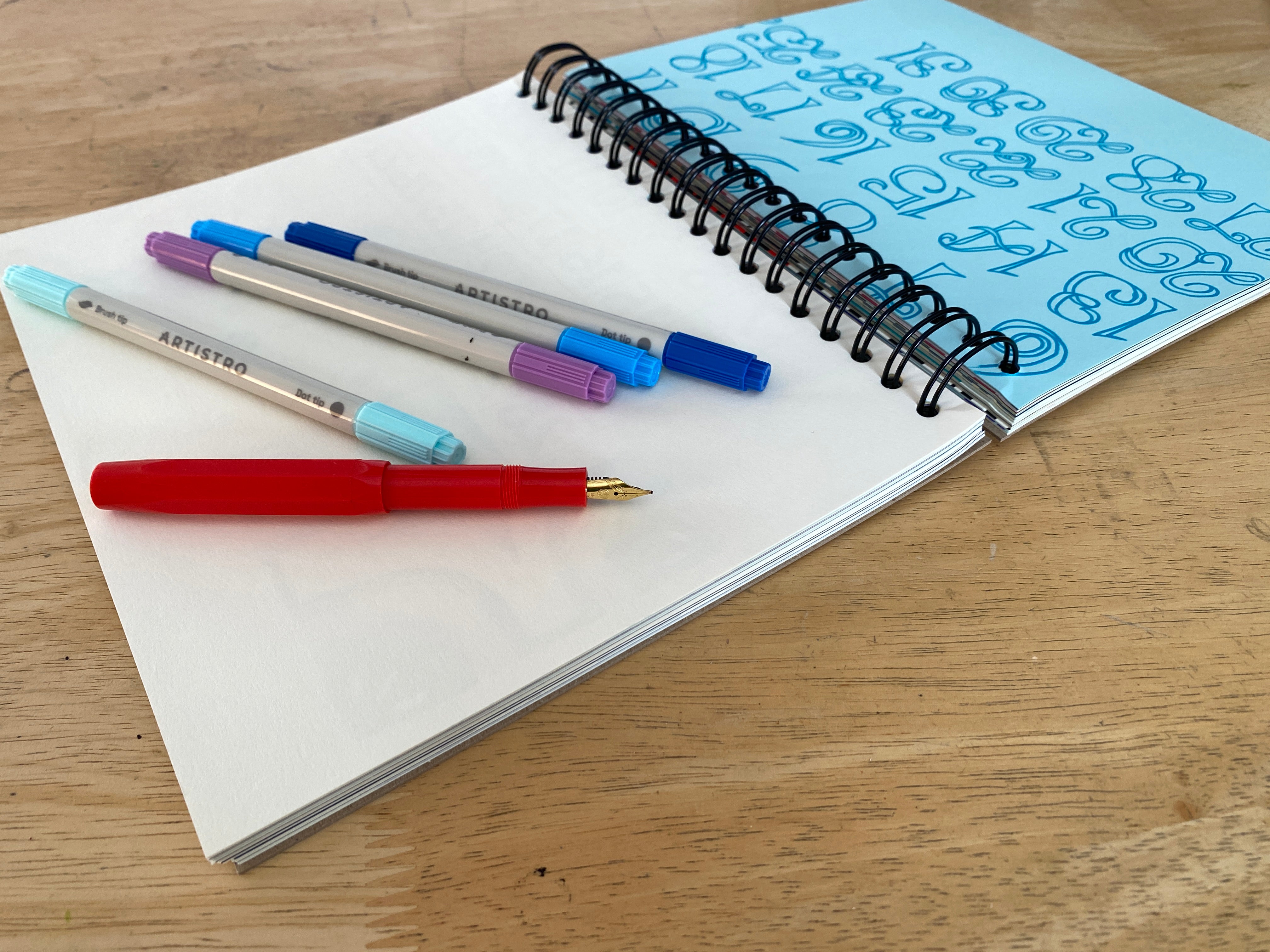 Open notebook with spiral binding on a wooden surface, containing colored pens.