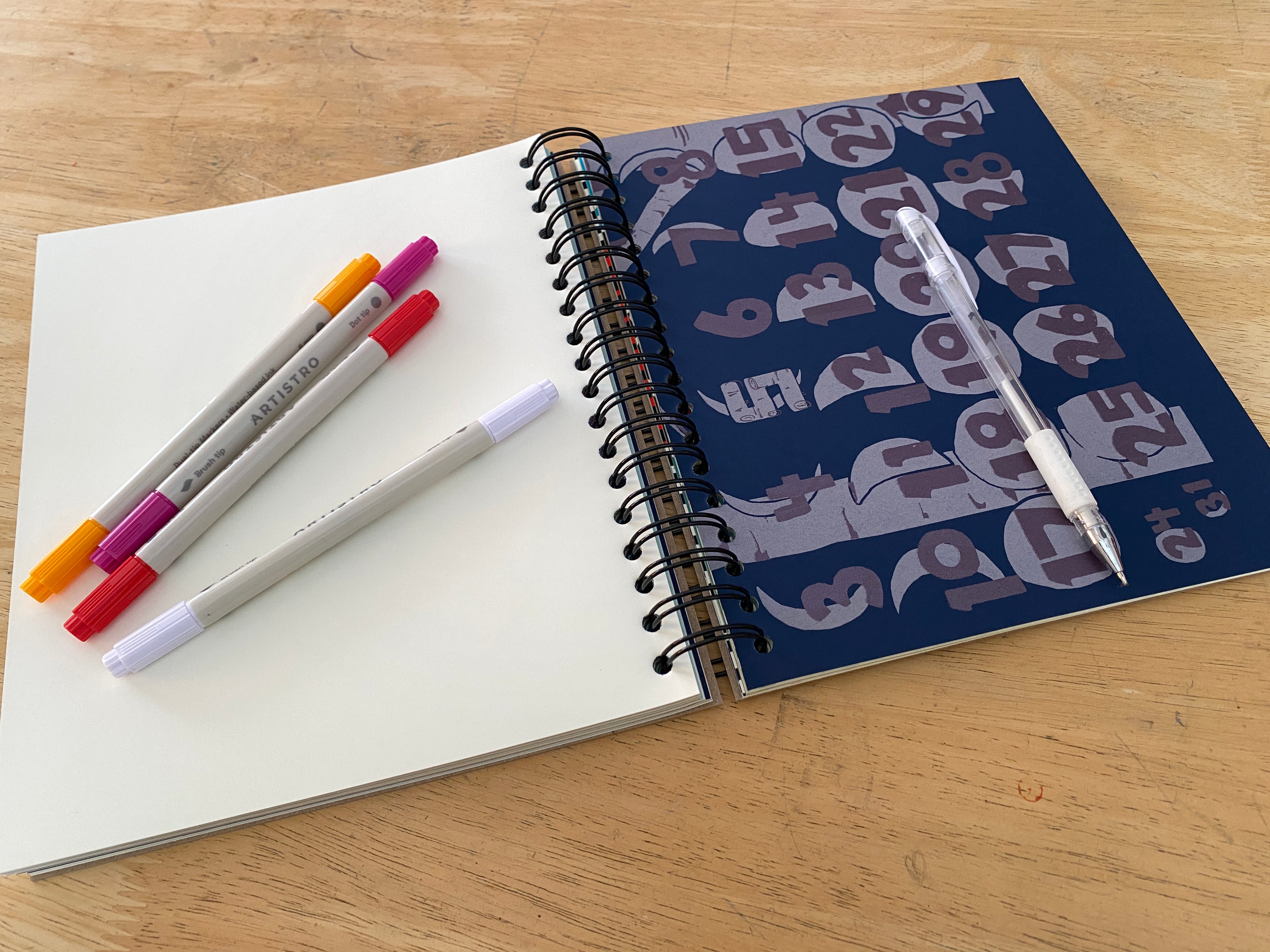 Open notebook with markers, a ruler, and a printed card on a wooden surface