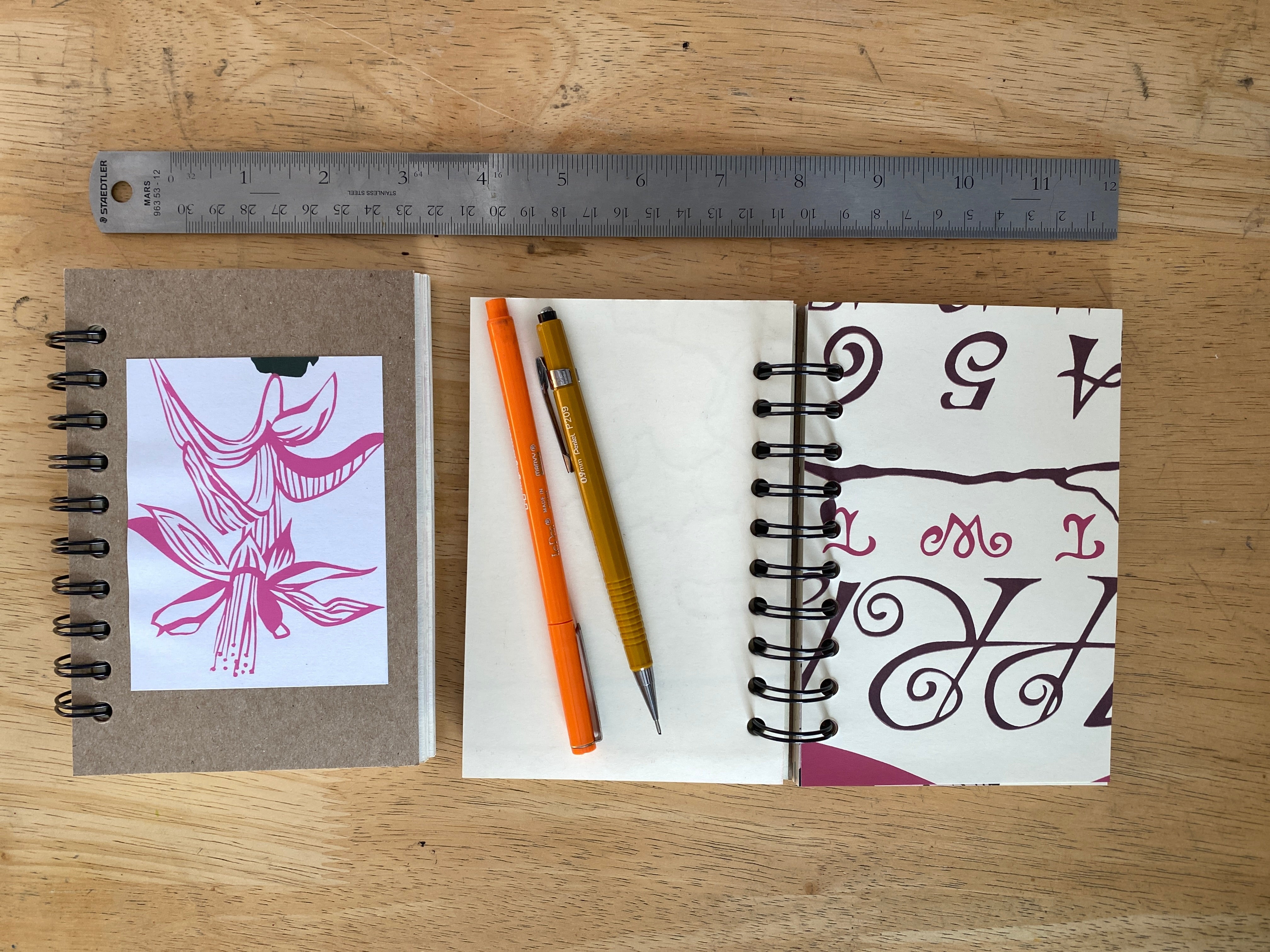 Two spiral-bound notebooks with printed designs, a pen, and a ruler on a wooden surface.