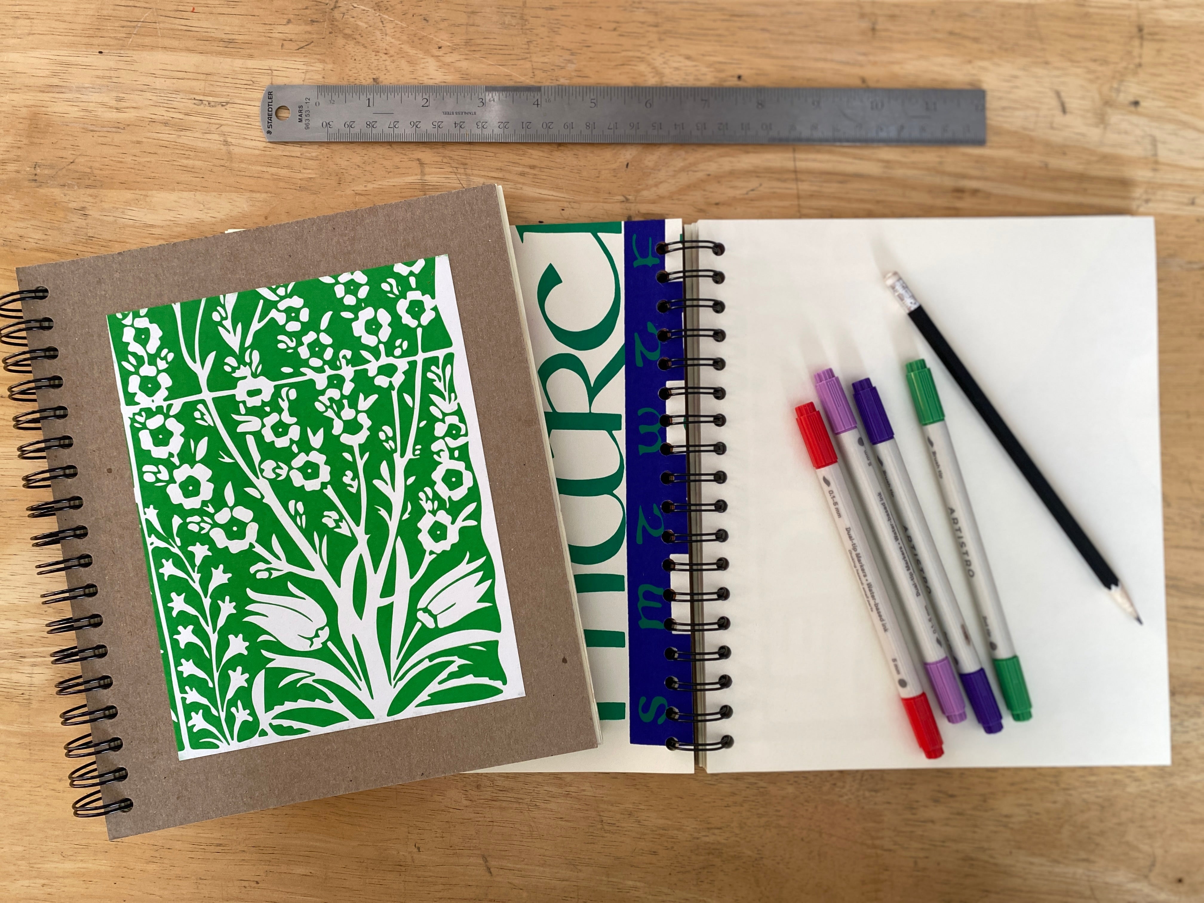Notebook with green floral design, markers, and a ruler on a wooden surface