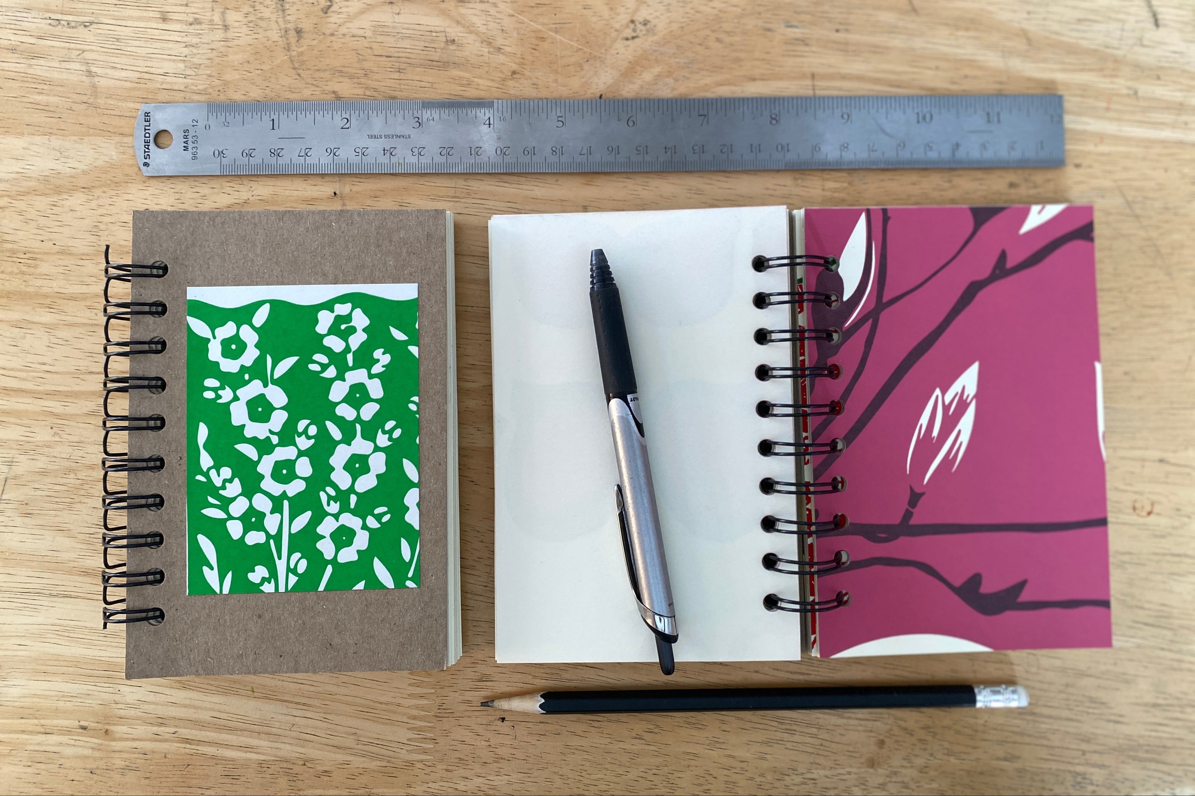 Two small notebooks with different designs on a wooden surface, accompanied by a pen and ruler.